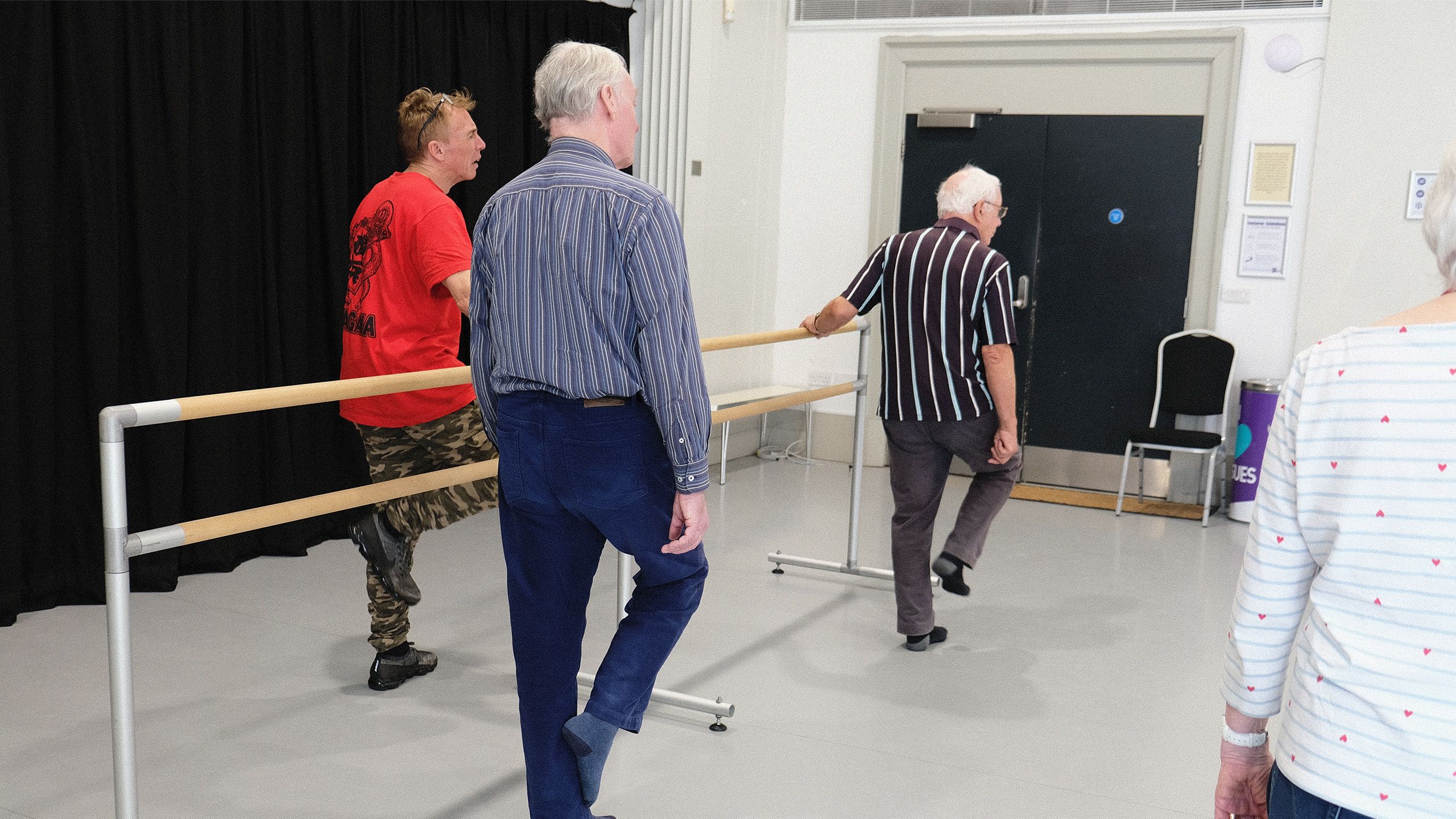 Three male attendees of Parkinson's class holding ballet bar and lifting their legs